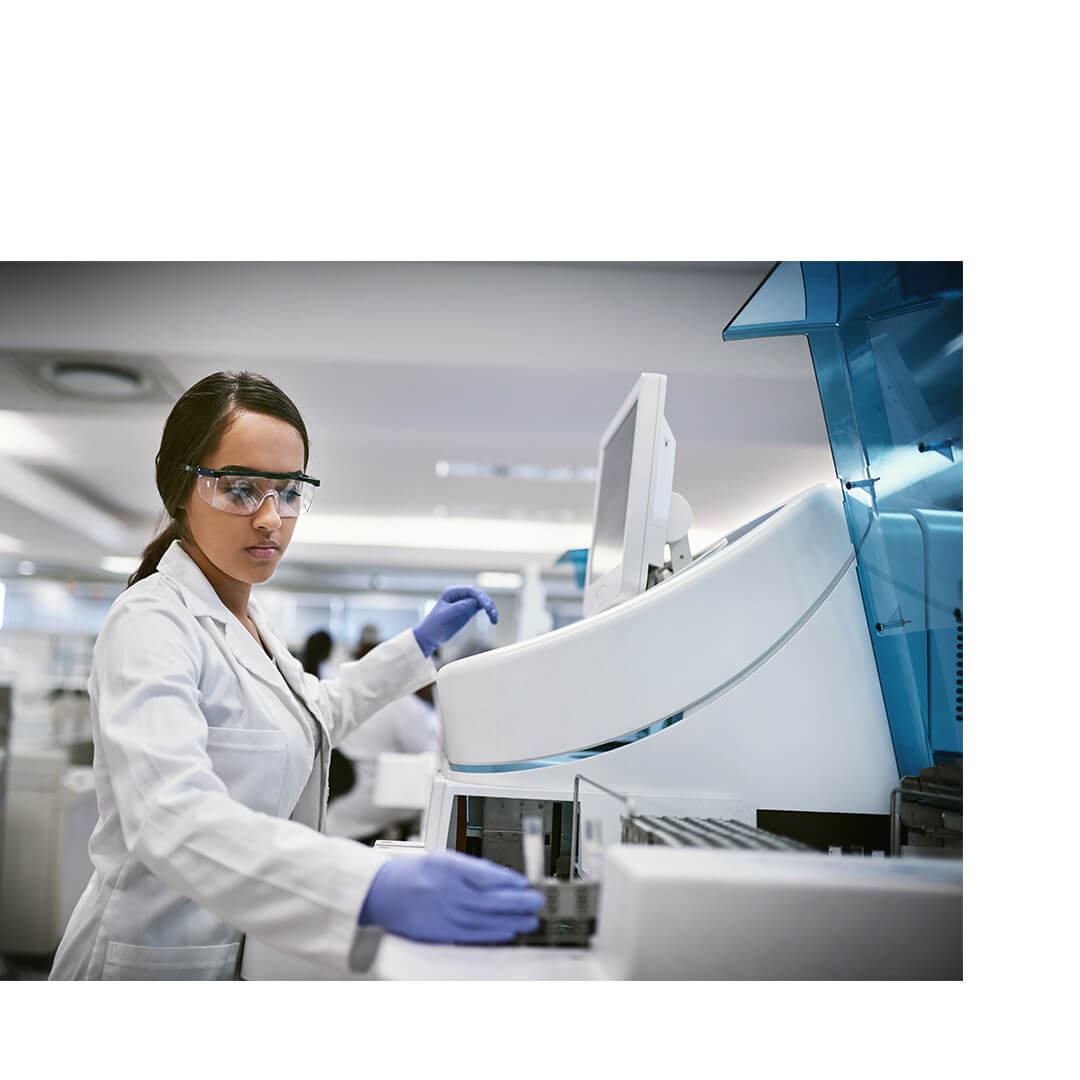 A female scientist in a lab coat and protective eyewear operates complex lab equipment in a bright research lab focused on cancer clinical trials