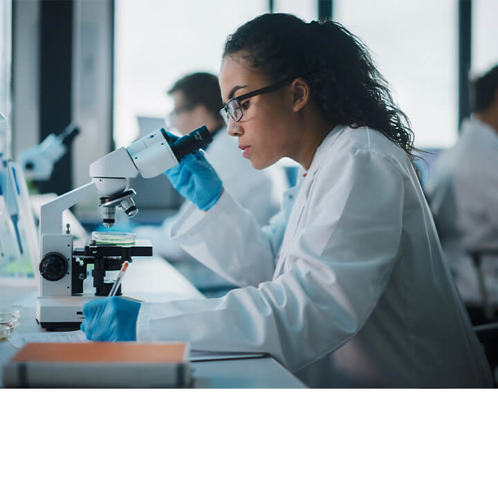 Female scientist wearing safety goggles and gloves operates a microscope in a cancer research lab