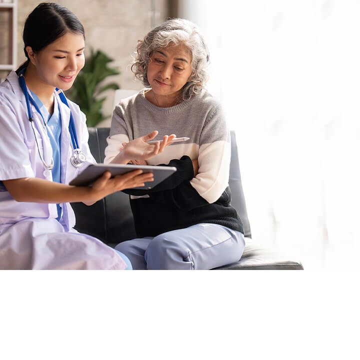 A nurse and an older adult patient sit together reviewing information on a tablet in a consultation room