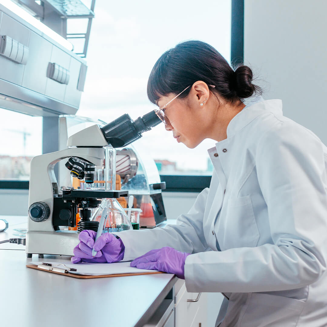 A woman in a lab coat examines a microscope, focused on her work in cancer research