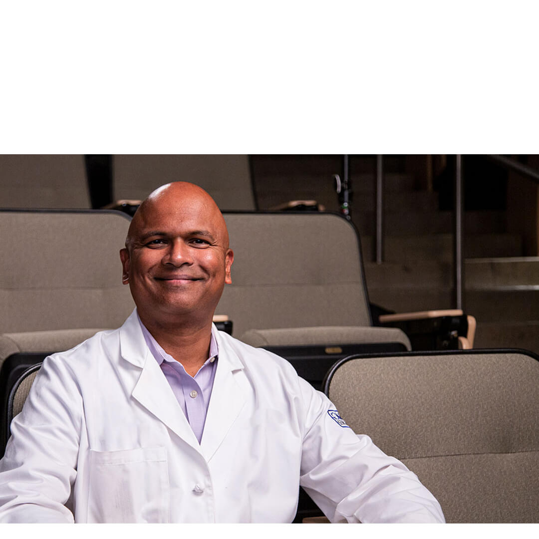 Dr. Sal Vasireddy, a smiling hematologist‑oncologist in a white coat, sits confidently in an auditorium-style clinic room, embodying compassionate cancer care